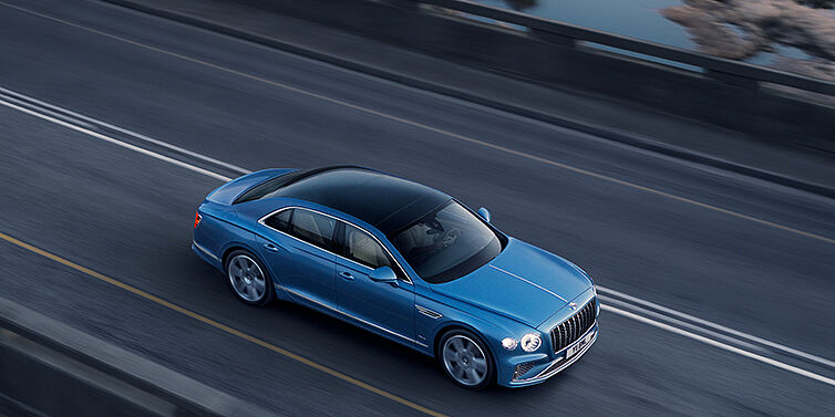 Bentley Geneve New Bentley Flying Spur Azure sedan in Light windsor blue paint by Mulliner, with 22 inch Azure wheels seen from overhead driving dynamically on a country road