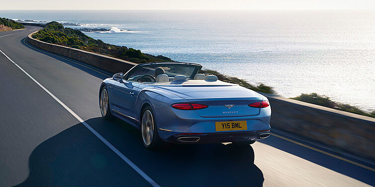 Bentley Geneve New Bentley Continental GTC Azure convertible rear view in Light Windsor blue paint with 22 inch ten spoke wheels driving dynamically on an ocean road in the sunshine